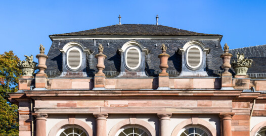 Schloss und Schlossgarten Schwetzingen - Garten - Nördlicher Zirkelbau - Westlicher Eck-Pavillon - Von Süden - Blick auf den oberen Teil der Südfassade sowie auf das Gebälk und den Schmuck (Vasen und Blumenkörbe) (Gesamter Bau: Länge Bogen ca. 175 Meter, Breite zwischen ca. 11 und 18 Meter, 51 Achsen, 5 Pavillons, 4 Zwischenbauten; Architekt: Alessandro Alessandro Galli da Bibiena (1686-1748), Bauleitung: zuerst Guillaume d’Hauberat (1680–1749), dann Nicolas de Pigage (1723-1796); Bauzeit: 1748-1750) (aufgenommen im Oktober 2024, am späten Vormittag)