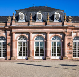 Schloss und Schlossgarten Schwetzingen - Garten - Nördlicher Zirkelbau - Mittlerer Pavillon - Von Südwest - Blick auf die Südwestfassade sowie auf das Gebälk und den Schmuck (Vasen und Blumenkörbe) (Gesamter Bau: Länge Bogen ca. 175 Meter, Breite zwischen ca. 11 und 18 Meter, 51 Achsen, 5 Pavillons, 4 Zwischenbauten; Architekt: Alessandro Alessandro Galli da Bibiena (1686-1748), Bauleitung: zuerst Guillaume d’Hauberat (1680–1749), dann Nicolas de Pigage (1723-1796); Bauzeit: 1748-1750) (aufgenommen im Oktober 2024, um die Mittagszeit)