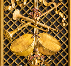 Schloss und Schlossgarten Schwetzingen - Garten - Schlosstheater - Von innen - Proszenium, Nordseite - Blick auf einen Teil der Trompeterloge mit Musikinstrumenten (Trompeten, ein Paar Pauken, Tamburin), einer Krone und einem Degen auf einem Gitterwerk sowie auf Girlanden, einem verschlungenen Band und weiteren Verzierungen (Gesamter Bau: Länge x Breite: ca. 62 x 35 Meter; Architekt: Nicolas de Pigage (1723-1796); Bauzeit: 1752-1753, Erweiterung 1762, Sanierung: 1937, Renovierung: 1952, Generalsanierung / Umbau 1971-1974, Restaurierung: 1999-2005) (aufgenommen im Dezember 2024, am späten Vormittag)