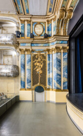 Schloss und Schlossgarten Schwetzingen - Garten - Schlosstheater - Von innen - Proszenium, Südseite - Von Norden - Blick von der Nordseite des Proszeniums auf die Südseite; der Orchestergraben ist auf halber Höhe angehoben; im unteren Teil der Südseite (vom Parkett bis zum Rang) die komposite Pilasterordnung; in der Mitte die Trompeterloge mit Musikinstrumenten (Flöten, Geige, Horn, Laute) und Notenblätter auf einem Gitterwerk sowie auf Girlanden, einem verschlungenen Band, einer Maske und weiteren Verzierungen, unten die Tür zum Orchestergraben; im oberen Teil der Südseite (Rang bis zur Decke) die korinthische Pilasterordnung; rechts oben der Architravbalken mit  Volutenkonsolen (Gesamter Bau: Länge x Breite: ca. 62 x 35 Meter; Architekt: Nicolas de Pigage (1723-1796); Bauzeit: 1752-1753, Erweiterung 1762, Sanierung: 1937, Renovierung: 1952, Generalsanierung / Umbau 1971-1974, Restaurierung: 1999-2005) (aufgenommen im Dezember 2024, am frühen Nachmittag)