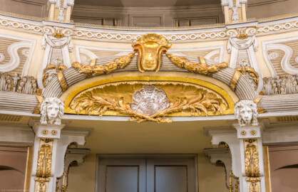 Schloss und Schlossgarten Schwetzingen - Garten - Schlosstheater - Von innen - Balkon, über dem Haupteingang zum Zuschauerraum - Von Westen - Blick vom Parkett auf eine stichbogige Lunette, darin eine Muschel und zwei davon ausgehende Blütengirlanden, mit kanneliertem Rundstab um den Stichbogen, oben eine vergoldete, plastisch geformte Agraffe mit zwei Laubwerkgirlanden, zu beiden Seiten der Lunette eine Löwenmaske (Gesamter Bau: Länge x Breite: ca. 62 x 35 Meter; Architekt: Nicolas de Pigage (1723-1796); Bauzeit: 1752-1753, Erweiterung 1762, Sanierung: 1937, Renovierung: 1952, Generalsanierung / Umbau 1971-1974, Restaurierung: 1999-2005) (aufgenommen im Dezember 2024, am frühen Nachmittag)