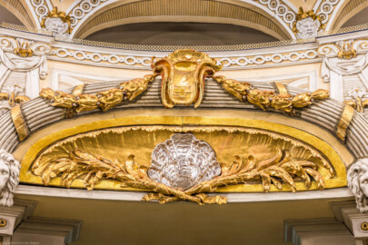 Schloss und Schlossgarten Schwetzingen - Garten - Schlosstheater - Von innen - Balkon, über dem Haupteingang zum Zuschauerraum - Von Westen - Blick vom Parkett auf eine stichbogige Lunette, darin eine Muschel und zwei davon ausgehende Blütengirlanden, mit kanneliertem Rundstab um den Stichbogen, oben eine vergoldete, plastisch geformte Agraffe mit zwei Laubwerkgirlanden, zu beiden Seiten der Lunette eine Löwenmaske (Gesamter Bau: Länge x Breite: ca. 62 x 35 Meter; Architekt: Nicolas de Pigage (1723-1796); Bauzeit: 1752-1753, Erweiterung 1762, Sanierung: 1937, Renovierung: 1952, Generalsanierung / Umbau 1971-1974, Restaurierung: 1999-2005) (aufgenommen im Dezember 2024, am frühen Nachmittag)