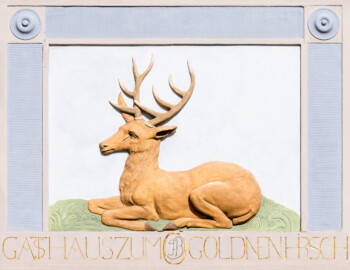 Schwetzingen - Schlossplatz, Nordseite - Palais Hirsch - Von außen - Von Süden - Hauszeichen über dem Portal des Palais Hirsch (Architekt: Alessandro Galli da Bibiena (1686-1748), Baujahr: 1748, Standort: am damals neu angelegten Marktplatz; Renovierung/Umbau: 1970er Jahre; ursprünglich erbaut im Auftrag des Kurfürsten Carl Theodor für seinen Berater und Beichtvater, den Jesuitenpfarrer Franz Joseph Seedorf (1691-1758); ab 1818: Nutzung als „Gasthaus zum Goldenen Hirsch“; heute: städtisches Gebäude für repräsentative Zwecke, Veranstaltungsort für Konferenzen, Tagungen, Seminare und Empfänge, ebenso Sitz der Forschungsstelle Geschichte der Südwestdeutschen Hofmusik der Heidelberger Akademie der Wissenschaften sowie Ausstellungsort des Kunstvereins Schwetzingen) (aufgenommen im März 2025, am späten Vormittag)