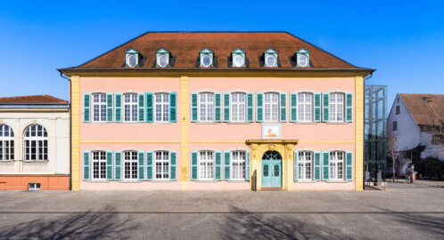 Schwetzingen - Schlossplatz, Nordseite - Palais Hirsch - Von außen - Von Süden - Blick vom Schlossplatz auf das Palais Hirsch (Architekt: Alessandro Galli da Bibiena (1686-1748), Baujahr: 1748, Standort: am damals neu angelegten Marktplatz; Renovierung/Umbau: 1970er Jahre; ursprünglich erbaut im Auftrag des Kurfürsten Carl Theodor für seinen Berater und Beichtvater, den Jesuitenpfarrer Franz Joseph Seedorf (1691-1758); ab 1818: Nutzung als „Gasthaus zum Goldenen Hirsch“; heute: städtisches Gebäude für repräsentative Zwecke, Veranstaltungsort für Konferenzen, Tagungen, Seminare und Empfänge, ebenso Sitz der Forschungsstelle Geschichte der Südwestdeutschen Hofmusik der Heidelberger Akademie der Wissenschaften sowie Ausstellungsort des Kunstvereins Schwetzingen) (aufgenommen im März 2025, am späten Vormittag)