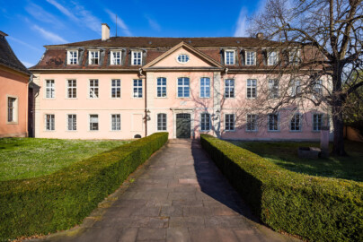 Schloss und Schlossgarten Schwetzingen - Garten - Gesandtenhaus (ehemaliges Prinzenhaus) / Amtsgericht - Von Südost - Blick von der Zeyherstraße auf die Südostfassade und das Portal des Gesandtenhauses; links die Rückseite des mittleren Pavillons des nördlichen Zirkelbaus; der Baum rechts ist eine Sommer-Linde (Tilia platyphyllos, Baum-Nr. 69) (Nutzung: Wohnungen für Angehörige der kurfürstlichen Familie, ausländische Besucher, Nicolas de Pigage (1723-1796), Friedrich Ludwig Skell (1750-1823), Johann Michael Zeyher (1770-1843), Sterbeort von Johann Peter Hebel (1760-1826), ab 1805 Amtsgericht) (Bau: Länge x Breite: ca. 40 x 17 Meter; Bauzeit: um 1723; Architekt: unbekannt) (aufgenommen im April 2025, am Vormittag)
