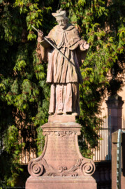 Schloss und Schlossgarten Schwetzingen - Schloss - Nahe dem Ehrenhof - Grünstreifen zwischen östlichem Ehrenhof und Leimbach - Nordostecke - Von Osten - Blick auf die Statue des Heiligen Nepomuk; Inschrift: "S. IOHANN VON NEPOMUCK / VON WENCELS WUTH BEKRIEGET / HAST AUFF DER PRAGER BRÜCK / DURCH SHWEIGEN OBSIEGET / ZUM SIEGES DENCKMAL HIER / THUT SHWETZINGEN AUFRICHTEN / DEIN BILDNIS UND SICH DIR / ZUR DIENERSCHAFT VERPFLICHTET / 1751" (zu Johannes (von) Nepomuk (um 1350-1393): böhmischer Priester und Märtyrer, 1729 heiliggesprochen, seit 1732 zweiter Ordenspatron der Jesuiten,  Brückenheiliger und Patron des Beichtgeheimnisses) (Statue ist eine Kopie, angefertigt 1927 und 1968 an diesem Ort aufgestellt; Original im Museum der Stadt Schwetzingen; Original wohl aus der Werkstatt des Hofbildhauers Paul Johann Egell (1691-1752), Datierung 1751) (aufgenommen im April 2025, am späten Vormittag)