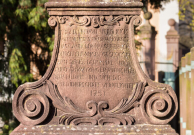 Schloss und Schlossgarten Schwetzingen - Schloss - Nahe dem Ehrenhof - Grünstreifen zwischen östlichem Ehrenhof und Leimbach - Nordostecke - Von Osten - Blick auf den Sockel der Statue des Heiligen Nepomuk; Inschrift: "S. IOHANN VON NEPOMUCK / VON WENCELS WUTH BEKRIEGET / HAST AUFF DER PRAGER BRÜCK / DURCH SHWEIGEN OBSIEGET / ZUM SIEGES DENCKMAL HIER / THUT SHWETZINGEN AUFRICHTEN / DEIN BILDNIS UND SICH DIR / ZUR DIENERSCHAFT VERPFLICHTET / 1751" (zu Johannes (von) Nepomuk (um 1350-1393): böhmischer Priester und Märtyrer, 1729 heiliggesprochen, seit 1732 zweiter Ordenspatron der Jesuiten,  Brückenheiliger und Patron des Beichtgeheimnisses) (Statue ist eine Kopie, angefertigt 1927 und 1968 an diesem Ort aufgestellt; Original im Museum der Stadt Schwetzingen; Original wohl aus der Werkstatt des Hofbildhauers Paul Johann Egell (1691-1752), Datierung 1751) (aufgenommen im April 2025, am späten Vormittag)