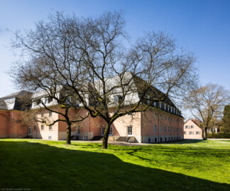 Schloss und Schlossgarten Schwetzingen - Garten - Schlosstheater - Von Nordost - Blick von der Nordwestecke des Gesandtenhauses nach Südwest auf den Gesandtenhausgarten sowie auf die Ostfassade (links) und Nordfassade (rechts) des Theaterbaus; die erste Treppe ganz rechts führt in das nördliche Treppenhaus, die Treppe links davon führt in das Foyer, die Treppe links davon führt in das Vestibül, die vierte Treppe ganz links führt in das südliche Treppenhaus; ganz links die Rückseite des nördlichen Zirkelbaus; der Bau zwischen Theater und dem rechten Baum ist das Fuhrmannshaus; in der Bildmitte zwei Schnurbäume und rechts ein weiterer Schnurbaum  (Sophora japonica, links Baum-Nr. 72 und 73, rechts Baum-Nr. 85) (Gesamter Bau: Länge x Breite: ca. 62 x 35 Meter; Architekt: Nicolas de Pigage (1723-1796); Bauzeit: 1752-1753, Erweiterung 1762, Sanierung: 1937, Renovierung: 1952, Generalsanierung / Umbau 1971-1974, Restaurierung: 1999-2005) (aufgenommen im April 2025, am frühen Nachmittag)