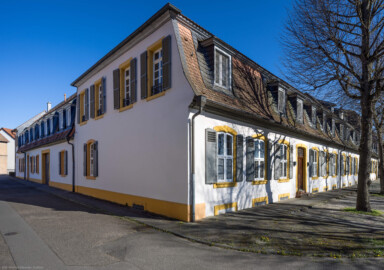 Schloss und Schlossgarten Schwetzingen - Sonstige Bauten - Palais Ysenburg (Forsthausstraße 7 und 9) - Von Nordwest - Blick vom großen Tor / der Durchfahrt zum Schlossgarten in der Forsthausstraße auf das Palais Ysenburg; links die Nordseite, rechts die Westseite (ehemalige Bewohner: Mitglieder des kurfürstlichen Hofstaats, Oberhofgärter Theodor van Wynder († 1777), bis 1778 Oberstjägermeister Graf Friedrich Wilhelm von Ysenburg-Birstein (1730-1804) und Ehefrau Caroline Gräfin von Parkstein (1762-1816), eine uneheliche Tochter von Kurfürst Carl Theodor, Oberstjägermeister Johann Theodor Maria Reichsgraf v. Waldkirch (1720–1802), Oberforstmeister Friedrich Heinrich Georg Freiherr von Drais (1758-1833), ab 1864 der Industrielle Gustav Bassermann und Ehefrau Clementine; nach 1918 Kauf durch Stadt Schwetzingen, seit 1997 in Privatbesitz) (Bau: L-formig; Länge x Breite: Nordflügel ca. 34 x 7 Meter, Westflügel ca. 21 x 13 Meter, Westflügel mit Anbau bis zum Alten Forstamt ca. 48 x 10 Meter; Geschosse: 2 (inkl. Mansardgeschoss); Bauzeiten: 1760, Verbindung zum Alten Forstamt 1775, erweitert Ende des 19. Jh., restauriert 1997; Architekt: wohl Franz Wilhelm Rabaliatti (1714–1782)) (aufgenommen im April 2025, am späten Nachmittag)