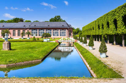 Schloss und Schlossgarten Schwetzingen - Garten - Orangeriegarten - Östlicher Kanal - Von Süden - Blick von der Südostecke des Orangeriegartens nach Norden auf den östlichen Kanal mit der Brücke; im Hintergrund der östliche Teil der Südfassade der Neuen Orangerie mit dem Glashaus (ganz rechts) (aufgenommen im April 2025, am Nachmittag)