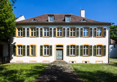 Schloss und Schlossgarten Schwetzingen - Sonstige Bauten - Altes Forstamt (Forsthausstraße 11) - Von Westen - Blick von der westlichen Gartenmauer nach Osten auf die Westfassade und den Haupteingang zum Forstamt; rechts im Hintergrund die Westseite der Remise (Bau: Länge x Breite: ca. 22 x 14 Meter; Geschosse: 2; Fensterachsen: 7; Bauzeit: 1760; erbaut als Haus des kurfürstlichen Oberjägermeisters; Verbindung mit dem Palais Ysenburg: 1775; Architekt: unbekannt) (aufgenommen im April 2025, am Nachmittag)