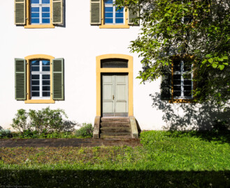 Schloss und Schlossgarten Schwetzingen - Sonstige Bauten - Altes Forstamt (Forsthausstraße 11) - Von Osten - Blick von der östlichen Gartenmauer nach Westen auf die Ostfassade und den Osteingang zum Forstamt; der Baum rechts ist eine Echte Walnuss (Juglans regia) (Bau: Länge x Breite: ca. 22 x 14 Meter; Geschosse: 2; Fensterachsen: 7; Bauzeit: 1760; erbaut als Haus des kurfürstlichen Oberjägermeisters; Verbindung mit dem Palais Ysenburg: 1775; Architekt: unbekannt) (aufgenommen im April 2025, am Vormittag)