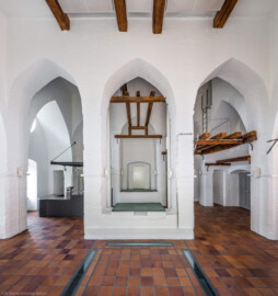 Schloss und Schlossgarten Schwetzingen - Sonstige Bauten - Oberes Wasserwerk - Wasserturm - Von innen - Obergeschoss / Cafeteria / Kantine - Von Süden - Zentraler Blick vom südlichen Fenster nach Norden in den Raum; Wände und mittige Pfeiler sind "gotisch" bzw. spitzbogenartig gestaltet; links die Theke der Cafeteria, rechts eine kleine Zwischenebene, die zum Dachstuhl über zwei Eisenleitern führt; in der Bildmitte eine Tür, die zum Dachstuhl des sog. Reißbodens führt; rechts und links der mittleren Pfeiler zwei der vier Leitungen (Steigleitungen? / Fallleitungen?) von der Brunnenstube zu den Wasserbehältern im Dachgeschoss; eiserne Anker in den Wänden und Pfeilern und Holzbalken dienten wohl als Zwischenebenen zur Lagerung (Gesamte Anlage Brunnenstube, Pumpenstube, Räderwerk, Wasserturm und Kühlhaus): Länge x Breite: ca. 30 x 29 Meter, Wasserturm: Länge x Breite: ca. 14 x 14 Meter, Firsthöhe ca. 24 Meter; Bauzeit Anlage: um 1774, Gesamtinstandsetzung: 1994–2000; Architekt: Nicolas de Pigage, Brunnenmeister: Thomas Breuer) (aufgenommen im Juni 2025, am frühen Nachmittag)