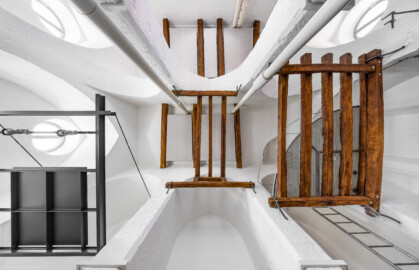 Schloss und Schlossgarten Schwetzingen - Sonstige Bauten - Oberes Wasserwerk - Wasserturm - Von innen - Obergeschoss / Cafeteria / Kantine - Nach oben - Blick vom Zentrum der nördlichen Fensterachse (dem Zentrum des nördlichen Teils des Raumes) nach oben; links ist Westen, unten ist Norden; Wände und mittige Pfeiler sind "gotisch" bzw. spitzbogenartig gestaltet; das Stahlgebilde links ist eine Art abgehängte "Decke" über der Theke; das Gitter aus Balken rechts ist eine kleine Zwischenebene zum Aufstieg in den Dachstuhl über Eisenleitern; oben rechts und links parallel zu den Pfeilern zwei Leitungen (Steigleitungen? / Fallleitungen?) von der Brunnenstube zu den Wasserbehältern im Dachgeschoss; eiserne Anker in den Pfeilern und Holzbalken dienten wohl als Zwischenebenen zur Lagerung (Gesamte Anlage Brunnenstube, Pumpenstube, Räderwerk, Wasserturm und Kühlhaus): Länge x Breite: ca. 30 x 29 Meter, Wasserturm: Länge x Breite: ca. 14 x 14 Meter, Firsthöhe ca. 24 Meter; Bauzeit Anlage: um 1774, Gesamtinstandsetzung: 1994–2000; Architekt: Nicolas de Pigage, Brunnenmeister: Thomas Breuer) (aufgenommen im Juni 2025, am Nachmittag)