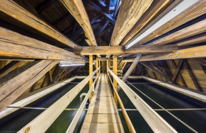 Schloss und Schlossgarten Schwetzingen - Sonstige Bauten - Oberes Wasserwerk - Wasserturm - Von innen - Dachstuhl - Von Norden - Zentraler Blick vom nördlichen Ende des Laufstegs über das Wasserbecken nach Süden in den Dachstuhl, auf den Laufsteg und das auf Wasserbecken; links etwas unterhalb der Bildmitte das Ende der Steigleitung mit dem U-förmigen Wasserzulauf; geschätzte Abmessungen des Behälters: Länge x Breite x Höhe 8 x 8 x 3 Meter, Fassungsvermögen ca. 170 Kubikmeter, Höhe über Boden ca. 17 Meter (Gesamte Anlage Brunnenstube, Pumpenstube, Räderwerk, Wasserturm und Kühlhaus): Länge x Breite: ca. 30 x 29 Meter, Wasserturm: Länge x Breite: ca. 14 x 14 Meter, Firsthöhe ca. 24 Meter; Bauzeit Anlage: um 1774, Gesamtinstandsetzung: 1994–2000; Architekt: Nicolas de Pigage, Brunnenmeister: Thomas Breuer) (aufgenommen im Juni 2025, am frühen Nachmittag)