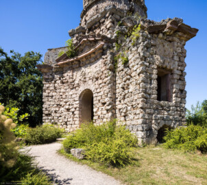 Schloss und Schlossgarten Schwetzingen - Garten - Merkurtempel - Von außen - Von Nordost - Blick auf die breite konkave Ostseite (links) und die schmale Nordseite (rechts) des Merkurtempels, als Ruine gebaut; das Mauerwerk besteht aus Tuffsteinquadern, die Gesimse aus Sandsteinblöcken, die Reliefs aus wetterfestem Marmorstuck; das Feld links zeigt Merkur als Befreier des Prometheus (nach der antiken Sage war aber Herakles / Herkules der Befreier des Prometheus), das Feld rechts zeigt ein Bukranion mit einer Girlande über den Hörnern; links der östliche rundbogige Eingang über wenige Treppenstufen, darüber ein Tympanon (darin ehemals geflügelte Hermesköpfe), rechts mittig ein Fenster eines Kabinetts, rechts unten ein Rundbogenfenster zur Beleuchtung des Untergeschosses (Architekt: Nicolas de Pigage (1723-1796); Bauzeit: 1784-1792, Restaurierung: 2008–2013; Abmessungen: Grundfläche des Sechsecks Länge x Breite ca. 14 x 12 Meter, Länge konkave Portalseiten ca. 10 Meter, Tiefe Kabinettseiten ca. 3,5 Meter, Durchmesser Tambour ca. 6,5 Meter, Höhe ca. 25 Meter, 4 Geschosse) (aufgenommen im Juni 2025, am frühen Nachmittag)