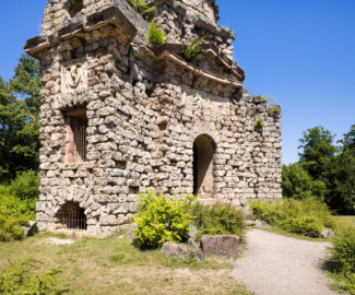 Schloss und Schlossgarten Schwetzingen - Garten - Merkurtempel - Von außen - Von Suedost - Blick auf die schmale Südseite (links) und die breite konkave Ostseite (rechts) des Merkurtempels, als Ruine gebaut; das Mauerwerk besteht aus Tuffsteinquadern, die Gesimse aus Sandsteinblöcken, die Reliefs aus wetterfestem Marmorstuck; das Feld links zeigt ein Bukranion mit einer Girlande über den Hörnern, das Feld rechts Merkur als Befreier des Prometheus (nach der antiken Sage war aber Herakles / Herkules der Befreier des Prometheus); links mittig ein Fenster des Treppenaufgangs, links unten ein Rundbogenfenster zur Beleuchtung des Untergeschosses, rechts der östliche rundbogige Eingang über wenige Treppenstufen, darüber ein Tympanon (darin ehemals geflügelte Hermesköpfe) (Architekt: Nicolas de Pigage (1723-1796); Bauzeit: 1784-1792, Restaurierung: 2008–2013; Abmessungen: Grundfläche des Sechsecks Länge x Breite ca. 14 x 12 Meter, Länge konkave Portalseiten ca. 10 Meter, Tiefe Kabinettseiten ca. 3,5 Meter, Durchmesser Tambour ca. 6,5 Meter, Höhe ca. 25 Meter, 4 Geschosse) (aufgenommen im Juni 2025, am frühen Nachmittag) Schloss und Schlossgarten Schwetzingen - Garten - Merkurtempel - Von außen - Von Suedost - Blick auf die schmale Südseite (links) und die breite konkave Ostseite (rechts) des Merkurtempels, als Ruine gebaut; das Mauerwerk besteht aus Tuffsteinquadern, die Gesimse aus Sandsteinblöcken, die Reliefs aus wetterfestem Marmorstuck; das Feld links zeigt ein Bukranion mit einer Girlande über den Hörnern, das Feld rechts Merkur als Befreier des Prometheus (nach der antiken Sage war aber Herakles / Herkules der Befreier des Prometheus); links mittig ein Fenster des Treppenaufgangs, links unten ein Rundbogenfenster zur Beleuchtung des Untergeschosses, rechts der östliche rundbogige Eingang über wenige Treppenstufen, darüber ein Tympanon (darin ehemals geflügelte Hermesköpfe) (Architekt: Nicolas de Pigage (1723-1796); Bauzeit: 1784-1792, Restaurierung: 2008–2013; Abmessungen: Grundfläche des Sechsecks Länge x Breite ca. 14 x 12 Meter, Länge konkave Portalseiten ca. 10 Meter, Tiefe Kabinettseiten ca. 3,5 Meter, Durchmesser Tambour ca. 6,5 Meter, Höhe ca. 25 Meter, 4 Geschosse) (aufgenommen im Juni 2025, am frühen Nachmittag)