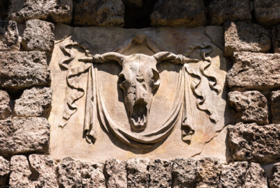 Schloss und Schlossgarten Schwetzingen - Garten - Merkurtempel - Südseite - Von außen - Von Süden - Blick auf ein Feld, oben an der Südseite des als Ruine gebauten Merkurtempels; das Stuckrelief zeigt ein Bukranion mit einer Girlande über den Hörnern (Bildhauer unklar, evtl. Giuseppe Antonio Pozzi (1732–1811), wohl nicht Franz Conrad Linck (1730-1793)) (Architekt: Nicolas de Pigage (1723-1796); Bauzeit: 1784-1792, Restaurierung: 2008–2013; Abmessungen: Grundfläche des Sechsecks Länge x Breite ca. 14 x 12 Meter, Länge konkave Portalseiten ca. 10 Meter, Tiefe Kabinettseiten ca. 3,5 Meter, Durchmesser Tambour ca. 6,5 Meter, Höhe ca. 25 Meter, 4 Geschosse) (aufgenommen im Juni 2025, um die Mittagszeit) Schloss und Schlossgarten Schwetzingen - Garten - Merkurtempel - Südseite - Von außen - Von Süden - Blick auf ein Feld, oben an der Südseite des als Ruine gebauten Merkurtempels; das Stuckrelief zeigt ein Bukranion mit einer Girlande über den Hörnern (Bildhauer unklar, evtl. Giuseppe Antonio Pozzi (1732–1811), wohl nicht Franz Conrad Linck (1730-1793)) (Architekt: Nicolas de Pigage (1723-1796); Bauzeit: 1784-1792, Restaurierung: 2008–2013; Abmessungen: Grundfläche des Sechsecks Länge x Breite ca. 14 x 12 Meter, Länge konkave Portalseiten ca. 10 Meter, Tiefe Kabinettseiten ca. 3,5 Meter, Durchmesser Tambour ca. 6,5 Meter, Höhe ca. 25 Meter, 4 Geschosse) (aufgenommen im Juni 2025, um die Mittagszeit)
