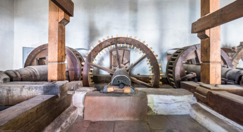 Schloss und Schlossgarten Schwetzingen - Sonstige Bauten - Oberes Wasserwerk - Pumpenstube - Von innen - Räder und Pumpen - Von Norden - Blick auf das Holzrad im Inneren der Stube, das mit der östlichen Welle / dem östlichen Wasserrad verbunden ist;  Holzrad: mit Holzzähnen im Radreifen, Durchmesser ca. 2,6 Meter; Welle im Holzrad: Durchmesser ca. 0,5 Meter, Länge ca. 6,5 Meter; das südliche Ende der Welle lagert in einer Nische an der Nordwand des Kellereigebäudes (im Hintergrund hinter dem Gitter etwas zu sehen); das mittige Holzrad greift links in ein weiteres Holzrad ein, das mit der Kurbelwelle der östlichen Pumpen verbunden ist; linkes Holzrad: Durchmesser ca. 1,8 Meter, mit zwei Radreifen, verbunden im Abstand von ca. 70 Zentimeter mit hölzernen Rundstäbe; das Holzrad rechts der Bildmitte ist mit der Kurbelwelle der westlichen Pumpen verbunden; das (nur teilweise sichtbare) Holzrad ganz rechts ist mit dem westlichen Wasserrad im Leimbach verbunden (Gesamte Anlage Brunnenstube, Pumpenstube, Räderwerk, Wasserturm und Kühlhaus): Länge x Breite: ca. 30 x 29 Meter, Pumpenstube: Länge x Breite: ca. 13 x 9 Meter; Bauzeit Anlage: um 1774, Gesamtinstandsetzung: 1994–2000; Architekt: Nicolas de Pigage, Brunnenmeister: Thomas Breuer) (aufgenommen im Juli 2025, am späten Vormittag)