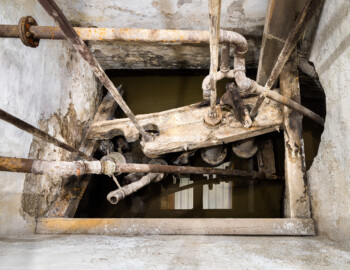 Schloss und Schlossgarten Schwetzingen - Sonstige Bauten - Oberes Wasserwerk - Pumpenstube - Von innen - Westliche Räder und Pumpen - Von Osten - Blick schräg nach unten in den westlichen Pumpenschacht (Länge x Breite x Tiefe ca. 2,5 x 2 x 4 Meter); rechts die Verbindung zu den Brunnen; im schräg verlaufenden Brett in der Mitte befinden sich die vier Hubkolbenpumpen aus Messing, darüber die vier Kolbenstangen mit einer Länge von ca. 2,9 Meter; unterhalb der linken beiden und oberhalb der rechten beiden Pumpen der Ansatz der Steigleitungen zu den Wasserbehältern im Wasserturm; unten im Bild sind Reflexion der hellen Bretter der Decke zu sehen (Gesamte Anlage Brunnenstube, Pumpenstube, Räderwerk, Wasserturm und Kühlhaus): Länge x Breite: ca. 30 x 29 Meter, Pumpenstube: Länge x Breite: ca. 13 x 9 Meter; Bauzeit Anlage: um 1774, Gesamtinstandsetzung: 1994–2000; Architekt: Nicolas de Pigage, Brunnenmeister: Thomas Breuer) (aufgenommen im Juli 2025, am späten Nachmittag)