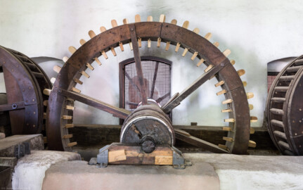 Schloss und Schlossgarten Schwetzingen - Sonstige Bauten - Oberes Wasserwerk - Pumpenstube - Von innen - Östliche Räder und Pumpen - Von Norden - Blick auf das Holzrad im Inneren der Stube, das mit dem östlichen Wasserrad verbunden ist;  Holzrad: mit Holzzähnen im Radreifen, Durchmesser ca. 2,6 Meter; Welle im Holzrad: Durchmesser ca. 0,5 Meter, Länge ca. 6,5 Meter; das südliche Ende der Welle lagert in einer Nische an der Nordwand des Kellereigebäudes (im Hintergrund hinter dem Gitter etwas zu sehen); das mittige Holzrad greift links in ein weiteres Holzrad ein, das mit der Kurbelwelle der östlichen Pumpen verbunden ist; linkes Holzrad: Durchmesser ca. 1,8 Meter, mit zwei Radreifen, verbunden im Abstand von ca. 70 Zentimeter mit hölzernen Rundstäbe (Gesamte Anlage Brunnenstube, Pumpenstube, Räderwerk, Wasserturm und Kühlhaus): Länge x Breite: ca. 30 x 29 Meter, Pumpenstube: Länge x Breite: ca. 13 x 9 Meter; Bauzeit Anlage: um 1774, Gesamtinstandsetzung: 1994–2000; Architekt: Nicolas de Pigage, Brunnenmeister: Thomas Breuer) (aufgenommen im Juli 2025, am späten Nachmittag)