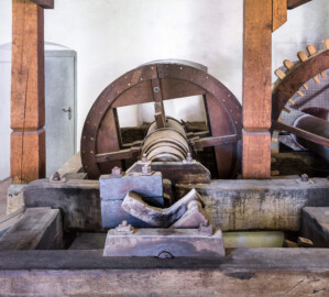 Schloss und Schlossgarten Schwetzingen - Sonstige Bauten - Oberes Wasserwerk - Pumpenstube - Von innen - Östliche Räder und Pumpen - Von Norden - Blick auf den östlichen Pumpenschacht, eingerahmt von vier Balken (vorne), darüber die Kurbelwelle (vorne oben), die mit der Kurbelwelle verbundene Holzwelle (Mitte) sowie das Holzrad (hinten); Pumpenschacht: Länge x Breite x Tiefe ca. 2,5 x 2 x 4 Meter; Kurbelwelle: aus Metall, für drei Kolbenstangen bzw. Kolben, die vorderste Kolbenstange ist ausgehängt und nach rechts gelegt; Welle: Durchmesser ca. 0,5 Meter; Holzrad: Durchmesser ca. 1,8 Meter, mit zwei Radreifen, verbunden im Abstand von ca. 70 Zentimeter mit hölzernen Rundstäbe; das mittige Holzrad greift rechts in ein weiteres Holzrad ein, das mit dem östlichen Wasserrad verbunden ist; rechtes Holzrad: mit Holzzähnen im Radreifen, Durchmesser ca. 2,6 Meter (Gesamte Anlage Brunnenstube, Pumpenstube, Räderwerk, Wasserturm und Kühlhaus): Länge x Breite: ca. 30 x 29 Meter, Pumpenstube: Länge x Breite: ca. 13 x 9 Meter; Bauzeit Anlage: um 1774, Gesamtinstandsetzung: 1994–2000; Architekt: Nicolas de Pigage, Brunnenmeister: Thomas Breuer) (aufgenommen im Juli 2025, am späten Nachmittag)