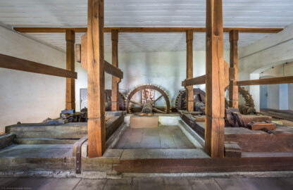 Schloss und Schlossgarten Schwetzingen - Sonstige Bauten - Oberes Wasserwerk - Pumpenstube - Von innen - Von Norden - Blick von der Nordwand des Pumpenhauses nach Süden in den Raum und auf die Pumpenanlage; hinter der Südwand befinden sich die beiden Wasserräder; in der Mitte und auf der linken Seite die Räder und Wellen der östlichen Pumpen, auf der rechten Seite die Räder und Wellen der westlichen Pumpen; die Tür ganz links führt auf einen Steg über den Leimbach und zur Schleuse vor den Wasserrädern; ganz rechts der Durchgang zwischen dem Pumpenhaus und dem Wasserturm (Gesamte Anlage Brunnenstube, Pumpenstube, Räderwerk, Wasserturm und Kühlhaus): Länge x Breite: ca. 30 x 29 Meter, Pumpenstube: Länge x Breite: ca. 13 x 9 Meter; Bauzeit Anlage: um 1774, Gesamtinstandsetzung: 1994–2000; Architekt: Nicolas de Pigage, Brunnenmeister: Thomas Breuer) (aufgenommen im Juli 2025, am Nachmittag)