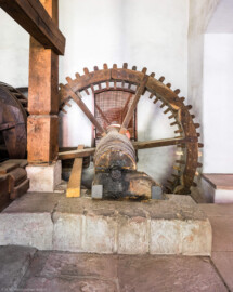 Schloss und Schlossgarten Schwetzingen - Sonstige Bauten - Oberes Wasserwerk - Pumpenstube - Von innen - Westliche Räder und Pumpen - Von Norden - Blick auf das Holzrad im Inneren der Stube, das mit dem westlichen Wasserrad verbunden ist; Holzrad: mit Holzzähnen im Radreifen, Durchmesser ca. 2,6 Meter; Welle im Holzrad: Durchmesser ca. 0,5 Meter, Länge ca. 6,5 Meter; das südliche Ende der Welle lagert in einer Nische an der Nordwand des Kellereigebäudes (im Hintergrund hinter dem Gitter etwas zu sehen); das mittige Holzrad greift links in ein weiteres Holzrad ein, das mit der Kurbelwelle der westlichen Pumpen verbunden ist; linkes Holzrad: Durchmesser ca. 1,8 Meter, mit zwei Radreifen, verbunden im Abstand von ca. 70 Zentimeter mit hölzernen Rundstäbe (Gesamte Anlage Brunnenstube, Pumpenstube, Räderwerk, Wasserturm und Kühlhaus): Länge x Breite: ca. 30 x 29 Meter, Pumpenstube: Länge x Breite: ca. 13 x 9 Meter; Bauzeit Anlage: um 1774, Gesamtinstandsetzung: 1994–2000; Architekt: Nicolas de Pigage, Brunnenmeister: Thomas Breuer) (aufgenommen im Juli 2025, am Nachmittag)