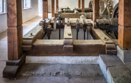 Schloss und Schlossgarten Schwetzingen - Sonstige Bauten - Oberes Wasserwerk - Pumpenstube - Von innen - Westliche Räder und Pumpen - Von Westen - Blick von der Westwand auf den westlichen Pumpenschacht, eingerahmt von vier Balken, darüber ein Querbalken in der Mitte (vorne), darüber die Kurbelwelle (Mitte), die mit der Kurbelwelle verbundene Holzwelle (rechts) sowie das Holzrad (ganz rechts); Pumpenschacht: Länge x Breite x Tiefe ca. 2,5 x 2 x 4 Meter; Kurbelwelle: aus Metall, für vier Kolbenstangen bzw. Kolben; Welle: Durchmesser ca. 0,5 Meter; Holzrad: Durchmesser ca. 1,8 Meter; im Hintergrund die Kurbelwelle der östlichen Pumpen (Gesamte Anlage Brunnenstube, Pumpenstube, Räderwerk, Wasserturm und Kühlhaus): Länge x Breite: ca. 30 x 29 Meter, Pumpenstube: Länge x Breite: ca. 13 x 9 Meter; Bauzeit Anlage: um 1774, Gesamtinstandsetzung: 1994–2000; Architekt: Nicolas de Pigage, Brunnenmeister: Thomas Breuer) (aufgenommen im Juli 2025, am späten Nachmittag)