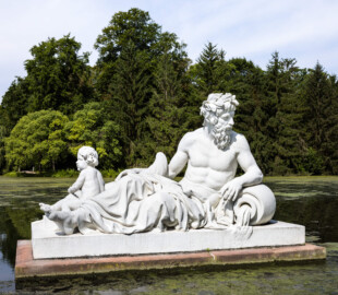 Schloss und Schlossgarten Schwetzingen - Garten - Um den Großen Weiher - Skulptur Rhein (rechts der Mitte des östlichen Ufers des Großen Weihers) - Von Osten - Blick auf die allegorische Kolossalfigur "Rhein"; dargestellt ist ein alter Flußgott, auf eine Urne gestützt lagernd, Kopf zur Seite gewendet, mit Kinn- und Oberlippenbart, mit einem Blattkranz auf dem Haar; links ein nackter Knabe, mit einem Fisch in der Hand; südlich der Figur befindet sich eine zweite Kolossalfigur "Donau"; ursprünglich waren auf der gegenüberliegenden Seite des Weihers noch die Figuren "Mosel" und "Maas" geplant; ursprünglicher Standort auf der Ufermauer, seit 1823 im umgestalteten Großen Weiher (Bildhauer: Peter Anton von Verschaffelt (1710-1793); Datierung: 1769 - 1779; Material: gelber Sandstein, weiß gefasst; Kopie; Original im Lapidarium) (aufgenommen im Juli 2025, am späten Vormittag)