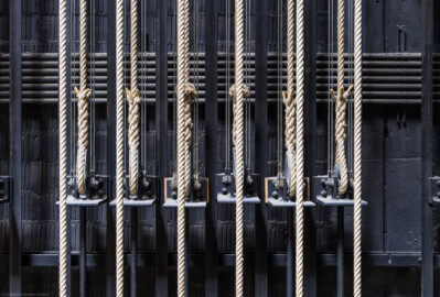 Schloss und Schlossgarten Schwetzingen - Garten - Schlosstheater - Von innen - Bühnenraum - Südliche Bühnenwand - Von Norden - Blick auf einige Handkonterzüge; die dicken Hanfseile sind die Kommandoseile, mit denen die Zugstangen (beladen z.B. mit Bühnenprospekt oder Scheinwerfern) in der Höhe verstellt werden können; das eine Ende eines Kommandoseils ist am Gegengewichtsschlitten (dem Winkel mit der Stange nach unten) befestigt; an diesem Schlitten sind auch die Stahlseile befestigt, die über Umlenkrollen die Last der Zugstange halten (Gesamter Bau: Länge x Breite: ca. 62 x 35 Meter; Architekt: Nicolas de Pigage (1723-1796); Bauzeit: 1752-1753, Erweiterung 1762, Sanierung: 1937, Renovierung: 1952, Generalsanierung / Umbau 1971-1974, Restaurierung: 1999-2005) (aufgenommen im Juli 2025, um die Mittagszeit) Schloss und Schlossgarten Schwetzingen - Garten - Schlosstheater - Von innen - Bühnenraum - Südliche Bühnenwand - Von Norden - Blick auf einige Handkonterzüge; die dicken Hanfseile sind die Kommandoseile, mit denen die Zugstangen (beladen z.B. mit Bühnenprospekt oder Scheinwerfern) in der Höhe verstellt werden können; das eine Ende eines Kommandoseils ist am Gegengewichtsschlitten (dem Winkel mit der Stange nach unten) befestigt; an diesem Schlitten sind auch die Stahlseile befestigt, die über Umlenkrollen die Last der Zugstange halten (Gesamter Bau: Länge x Breite: ca. 62 x 35 Meter; Architekt: Nicolas de Pigage (1723-1796); Bauzeit: 1752-1753, Erweiterung 1762, Sanierung: 1937, Renovierung: 1952, Generalsanierung / Umbau 1971-1974, Restaurierung: 1999-2005) (aufgenommen im Juli 2025, um die Mittagszeit)