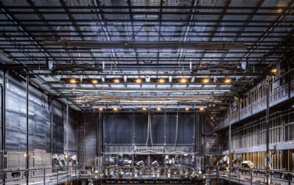 Schloss und Schlossgarten Schwetzingen - Garten - Schlosstheater - Von innen - Bühnenraum - Obermaschinerie - Von Westen - Blick von der westlichen Arbeitsgalerie nach Osten in den Bühnenraum und auf die Obermaschinerie; mittig unten die (in der Höhe einstellbare) begehbare Beleuchtungsbrücke mit umfangreicher Beleuchtungstechnik; darüber ein schwarzer Vorhang, hinter dem Vorhang der (nicht sichtbare) eiserne Vorhang; links und rechts unten die begehbare Arbeitsgalerie (Höhe ca. 8 Meter) mit Scheinwerfern auf dem Geländer; rechts die südliche Bühnenwand mit den ca. 55 Zügen zur Höhenverstellung der an Seilen aufgehängten horizontalen Zugstangen; ganz oben der begehbare Rollenboden / Schnürboden (Höhe ca. 13,5 Meter), unter dem sich Zugstangen für Bühnenprospekte, Beleuchtung etc. befinden; im Bild sind mehrere Zugstangen mit in beigen Leinwandtüchern eingewickelten Bühnenprospekten zu sehen (Gesamter Bau: Länge x Breite: ca. 62 x 35 Meter; Architekt: Nicolas de Pigage (1723-1796); Bauzeit: 1752-1753, Erweiterung 1762, Sanierung: 1937, Renovierung: 1952, Generalsanierung / Umbau 1971-1974, Restaurierung: 1999-2005) (aufgenommen im Juli 2025, am Nachmittag) Schloss und Schlossgarten Schwetzingen - Garten - Schlosstheater - Von innen - Bühnenraum - Obermaschinerie - Von Westen - Blick von der westlichen Arbeitsgalerie nach Osten in den Bühnenraum und auf die Obermaschinerie; mittig unten die (in der Höhe einstellbare) begehbare Beleuchtungsbrücke mit umfangreicher Beleuchtungstechnik; darüber ein schwarzer Vorhang, hinter dem Vorhang der (nicht sichtbare) eiserne Vorhang; links und rechts unten die begehbare Arbeitsgalerie (Höhe ca. 8 Meter) mit Scheinwerfern auf dem Geländer; rechts die südliche Bühnenwand mit den ca. 55 Zügen zur Höhenverstellung der an Seilen aufgehängten horizontalen Zugstangen; ganz oben der begehbare Rollenboden / Schnürboden (Höhe ca. 13,5 Meter), unter dem sich Zugstangen für Bühnenprospekte, Beleuchtung etc. befinden; im Bild sind mehrere Zugstangen mit in beigen Leinwandtüchern eingewickelten Bühnenprospekten zu sehen (Gesamter Bau: Länge x Breite: ca. 62 x 35 Meter; Architekt: Nicolas de Pigage (1723-1796); Bauzeit: 1752-1753, Erweiterung 1762, Sanierung: 1937, Renovierung: 1952, Generalsanierung / Umbau 1971-1974, Restaurierung: 1999-2005) (aufgenommen im Juli 2025, am Nachmittag)