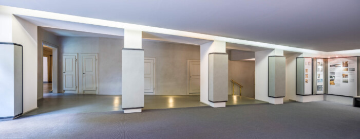 Schloss und Schlossgarten Schwetzingen - Garten - Schlosstheater - Von innen - Erdgeschoss, nördliches Foyer - Von Nordost - Blick nach Südwest in das nördliche Foyer im Erdgeschoss;  urprünglich befand sich die nördliche Mauer des Theaters an der Stelle, an der heute die drei Pfeiler stehen; in den 1950/1960er Jahren und vor allem beim großen Umbau 1971-1974 wurde das Schlosstheater nach Norden hin erweitert; originaler Bau und Anbau erkennbar am Boden: Holzboden bedeutet originaler Bau, Teppichboden bedeutet Anbau des 20. Jahrhunderts; hinter dem Durchgang auf der linken (östlichen) Seite liegt das östliche Foyer und das Vestibül; mittig die Zugänge zu den Logen im Parterre; rechts Tafeln einer Ausstellung zur Geschichte des Theaters (Gesamter Bau: Länge x Breite: ca. 62 x 35 Meter; Architekt: Nicolas de Pigage (1723-1796); Bauzeit: 1752-1753, Erweiterung 1762, Sanierung: 1937, Renovierung: 1952, Generalsanierung / Umbau 1971-1974, Restaurierung: 1999-2005) (aufgenommen im August 2025, um die Mittagszeit)