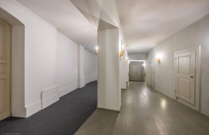 Schloss und Schlossgarten Schwetzingen - Garten - Schlosstheater - Von innen - Erdgeschoss, südliches Foyer - Von Osten - Blick von der Ostwand des Foyers nach Westen in das südliche Foyer; urprünglich befand sich die südliche Mauer des Theaters an der Stelle, an der heute die zwei Pfeiler stehen; beim großen Umbau 1971-1974 wurde das Schlosstheater zum nördlichen Zirkelbau (nach Süden / links) hin erweitert; originaler Bau und Anbau erkennbar am Boden: Holzboden bedeutet originaler Bau, Teppichboden bedeutet Anbau des 20. Jahrhunderts; rechts die Zugänge zu den Logen im Parterre; die große Tür links führt vom Foyer im nördlichen Zirkelbau in das südliche Foyer des Theaters; die Tür rechts der Bildmitte im Hintergrund führt zur Bühne, dem Orchestergraben und der Unterbühne; die (nicht sichtbare) Tür im Türrahmen rechts davon führt in das Parterre des Zuschauerraums (Gesamter Bau: Länge x Breite: ca. 62 x 35 Meter; Architekt: Nicolas de Pigage (1723-1796); Bauzeit: 1752-1753, Erweiterung 1762, Sanierung: 1937, Renovierung: 1952, Generalsanierung / Umbau 1971-1974, Restaurierung: 1999-2005) (aufgenommen im August 2025, am Nachmittag)