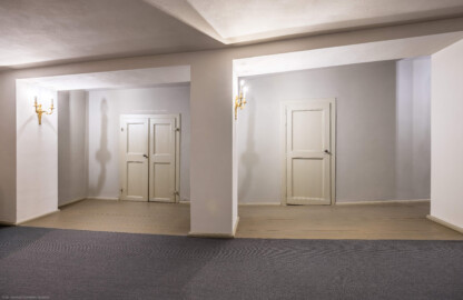 Schloss und Schlossgarten Schwetzingen - Garten - Schlosstheater - Von innen - 1. Obergeschoss (Balkon), südliches Foyer - Von Süden - Blick nach Nordost in das südliche Foyer auf die westlichen Türen zum Balkon; urprünglich befand sich die südliche Mauer des Theaters an der Stelle, an der heute die Pfeiler stehen; beim großen Umbau 1971-1974 wurde das Schlosstheater zum nördlichen Zirkelbau (nach Süden) hin erweitert; originaler Bau und Anbau erkennbar am Boden: Holzboden bedeutet originaler Bau, Teppichboden bedeutet Anbau des 20. Jahrhunderts (Gesamter Bau: Länge x Breite: ca. 62 x 35 Meter; Architekt: Nicolas de Pigage (1723-1796); Bauzeit: 1752-1753, Erweiterung 1762, Sanierung: 1937, Renovierung: 1952, Generalsanierung / Umbau 1971-1974, Restaurierung: 1999-2005) (aufgenommen im August 2025, am frühen Nachmittag)