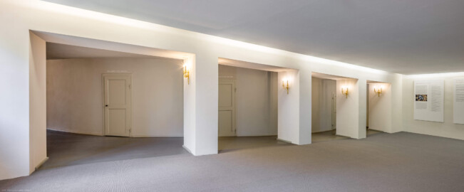 Schloss und Schlossgarten Schwetzingen - Garten - Schlosstheater - Von innen - 1. Obergeschoss (Balkon), nördliches Foyer - Von Nordost - Blick nach Südwest in das nördliche Foyer im 1. Obergeschoss;  urprünglich befand sich die nördliche Mauer des Theaters an der Stelle, an der heute die drei Pfeiler stehen; in den 1950/1960er Jahren und vor allem beim großen Umbau 1971-1974 wurde das Schlosstheater nach Norden hin erweitert; originaler Bau und Anbau erkennbar am Boden: Holzboden bedeutet originaler Bau, Teppichboden bedeutet Anbau des 20. Jahrhunderts; hinter dem Durchgang auf der linken (östlichen) Seite liegt das östliche Foyer; mittig die Zugänge zu den Rängen; rechts Tafeln einer Ausstellung zu den kurfürstlichen Opern und der kurfürstlichen Hofmusik (Gesamter Bau: Länge x Breite: ca. 62 x 35 Meter; Architekt: Nicolas de Pigage (1723-1796); Bauzeit: 1752-1753, Erweiterung 1762, Sanierung: 1937, Renovierung: 1952, Generalsanierung / Umbau 1971-1974, Restaurierung: 1999-2005) (aufgenommen im August 2025, am frühen Nachmittag)