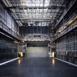 Schloss und Schlossgarten Schwetzingen - Garten - Schlosstheater - Von innen - Bühnenraum - Von Westen - Blick von der Hinterbühne nach Osten in den Bühnenraum und auf die Bühne; mittig die (in der Höhe einstellbare) begehbare Beleuchtungsbrücke mit umfangreicher Beleuchtungstechnik, darunter der geschlossene Eiserne Vorhang, dahinter (nicht sichtbar) der Zuschauerraum; rechts die südliche Bühnenwand mit den ca. 55 Zügen zur Höhenverstellung der an Stahlseilen aufgehängten horizontalen Zugstangen; links und rechts in halber Höhe die Arbeitsgalerie mit Scheinwerfern auf dem Geländer; rechts oberhalb der Arbeitsgalerie die Gewichtsgalerie zum Einhängen von Gewichten in die Gegengewichtsschlitten; links unten das geschlossene Seitenbühnentor, dahinter (nicht sichtbar) die Seitenbühne; links und rechts des Eisernen Vorhangs die begehbaren Portaltürme; ganz oben der begehbare Rollenboden / Schnürboden (Höhe ca. 13,5 Meter) mit Zugstangen unterhalb für Bühnenprospekte, Beleuchtung etc. (Gesamter Bau: Länge x Breite: ca. 62 x 35 Meter; Architekt: Nicolas de Pigage (1723-1796); Bauzeit: 1752-1753, Erweiterung 1762, Sanierung: 1937, Renovierung: 1952, Generalsanierung / Umbau 1971-1974, Restaurierung: 1999-2005) (aufgenommen im August 2025,am späten Vormittag) Schloss und Schlossgarten Schwetzingen - Garten - Schlosstheater - Von innen - Bühnenraum - Von Westen - Blick von der Hinterbühne nach Osten in den Bühnenraum und auf die Bühne; mittig die (in der Höhe einstellbare) begehbare Beleuchtungsbrücke mit umfangreicher Beleuchtungstechnik, darunter der geschlossene Eiserne Vorhang, dahinter (nicht sichtbar) der Zuschauerraum; rechts die südliche Bühnenwand mit den ca. 55 Zügen zur Höhenverstellung der an Stahlseilen aufgehängten horizontalen Zugstangen; links und rechts in halber Höhe die Arbeitsgalerie mit Scheinwerfern auf dem Geländer; rechts oberhalb der Arbeitsgalerie die Gewichtsgalerie zum Einhängen von Gewichten in die Gegengewichtsschlitten; links unten das geschlossene Seitenbühnentor, dahinter (nicht sichtbar) die Seitenbühne; links und rechts des Eisernen Vorhangs die begehbaren Portaltürme; ganz oben der begehbare Rollenboden / Schnürboden (Höhe ca. 13,5 Meter) mit Zugstangen unterhalb für Bühnenprospekte, Beleuchtung etc. (Gesamter Bau: Länge x Breite: ca. 62 x 35 Meter; Architekt: Nicolas de Pigage (1723-1796); Bauzeit: 1752-1753, Erweiterung 1762, Sanierung: 1937, Renovierung: 1952, Generalsanierung / Umbau 1971-1974, Restaurierung: 1999-2005) (aufgenommen im August 2025,am späten Vormittag)