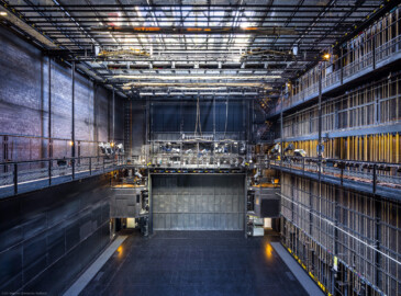 Schloss und Schlossgarten Schwetzingen - Garten - Schlosstheater - Von innen - Bühnenraum - Von Westen - Blick von der westlichen begehbaren Arbeitsgalerie in einer Höhe von ca. 8 Meter nach Osten in den Bühnenraum und auf die Bühne; mittig die (in der Höhe einstellbare) begehbare Beleuchtungsbrücke mit umfangreicher Beleuchtungstechnik, darunter der geschlossene Eiserne Vorhang, dahinter (nicht sichtbar) der Zuschauerraum; rechts die südliche Bühnenwand mit den ca. 55 Zügen zur Höhenverstellung der an Stahlseilen aufgehängten horizontalen Zugstangen; links und rechts in halber Höhe die Arbeitsgalerie mit Scheinwerfern auf dem Geländer; rechts oberhalb der Arbeitsgalerie die Gewichtsgalerie zum Einhängen von Gewichten in die Gegengewichtsschlitten; links unten das geschlossene Seitenbühnentor, dahinter (nicht sichtbar) die Seitenbühne; links und rechts des Eisernen Vorhangs die begehbaren Portaltürme; ganz oben der begehbare Rollenboden / Schnürboden (Höhe ca. 13,5 Meter) mit Zugstangen unterhalb für Bühnenprospekte, Beleuchtung etc. (Gesamter Bau: Länge x Breite: ca. 62 x 35 Meter; Architekt: Nicolas de Pigage (1723-1796); Bauzeit: 1752-1753, Erweiterung 1762, Sanierung: 1937, Renovierung: 1952, Generalsanierung / Umbau 1971-1974, Restaurierung: 1999-2005) (aufgenommen im August 2025, um die Mittagszeit) Schloss und Schlossgarten Schwetzingen - Garten - Schlosstheater - Von innen - Bühnenraum - Von Westen - Blick von der westlichen begehbaren Arbeitsgalerie in einer Höhe von ca. 8 Meter nach Osten in den Bühnenraum und auf die Bühne; mittig die (in der Höhe einstellbare) begehbare Beleuchtungsbrücke mit umfangreicher Beleuchtungstechnik, darunter der geschlossene Eiserne Vorhang, dahinter (nicht sichtbar) der Zuschauerraum; rechts die südliche Bühnenwand mit den ca. 55 Zügen zur Höhenverstellung der an Stahlseilen aufgehängten horizontalen Zugstangen; links und rechts in halber Höhe die Arbeitsgalerie mit Scheinwerfern auf dem Geländer; rechts oberhalb der Arbeitsgalerie die Gewichtsgalerie zum Einhängen von Gewichten in die Gegengewichtsschlitten; links unten das geschlossene Seitenbühnentor, dahinter (nicht sichtbar) die Seitenbühne; links und rechts des Eisernen Vorhangs die begehbaren Portaltürme; ganz oben der begehbare Rollenboden / Schnürboden (Höhe ca. 13,5 Meter) mit Zugstangen unterhalb für Bühnenprospekte, Beleuchtung etc. (Gesamter Bau: Länge x Breite: ca. 62 x 35 Meter; Architekt: Nicolas de Pigage (1723-1796); Bauzeit: 1752-1753, Erweiterung 1762, Sanierung: 1937, Renovierung: 1952, Generalsanierung / Umbau 1971-1974, Restaurierung: 1999-2005) (aufgenommen im August 2025, um die Mittagszeit)
