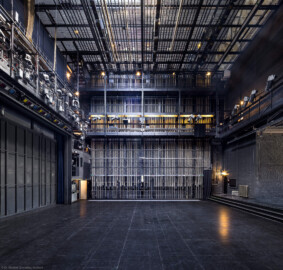 Schloss und Schlossgarten Schwetzingen - Garten - Schlosstheater - Von innen - Bühnenraum - Von Norden - Blick nach Süden auf die Bühne und in den Bühnenraum; links hinter dem Eisernen Vorhang der Zuschauerraum, rechts die Hinterbühne; in der Bildmitte die südliche Bühnenwand mit den ca. 55 Zügen zur Höhenverstellung der an Stahlseilen aufgehängten horizontalen Zugstangen; links in halber Höhe die (in der Höhe einstellbare) begehbare Beleuchtungsbrücke mit umfangreicher Beleuchtungstechnik; mittig und rechts in halber Höhe die begehbare Arbeitsgalerie (Höhe ca. 8 Meter) mit Scheinwerfern auf dem Geländer; mittig oberhalb der Arbeitsgalerie die Gewichtsgalerie zum Einhängen von Gewichten in die Gegengewichtsschlitten; ganz oben der begehbare Rollenboden / Schnürboden (Höhe ca. 13,5 Meter) mit Zugstangen unterhalb für Bühnenprospekte, Beleuchtung etc.; links der Wand mit den Zügen der südliche begehbare Portalturm (Gesamter Bau: Länge x Breite: ca. 62 x 35 Meter; Architekt: Nicolas de Pigage (1723-1796); Bauzeit: 1752-1753, Erweiterung 1762, Sanierung: 1937, Renovierung: 1952, Generalsanierung / Umbau 1971-1974, Restaurierung: 1999-2005) (aufgenommen im August 2025, um die Mittagszeit) Schloss und Schlossgarten Schwetzingen - Garten - Schlosstheater - Von innen - Bühnenraum - Von Norden - Blick nach Süden auf die Bühne und in den Bühnenraum; links hinter dem Eisernen Vorhang der Zuschauerraum, rechts die Hinterbühne; in der Bildmitte die südliche Bühnenwand mit den ca. 55 Zügen zur Höhenverstellung der an Stahlseilen aufgehängten horizontalen Zugstangen; links in halber Höhe die (in der Höhe einstellbare) begehbare Beleuchtungsbrücke mit umfangreicher Beleuchtungstechnik; mittig und rechts in halber Höhe die begehbare Arbeitsgalerie (Höhe ca. 8 Meter) mit Scheinwerfern auf dem Geländer; mittig oberhalb der Arbeitsgalerie die Gewichtsgalerie zum Einhängen von Gewichten in die Gegengewichtsschlitten; ganz oben der begehbare Rollenboden / Schnürboden (Höhe ca. 13,5 Meter) mit Zugstangen unterhalb für Bühnenprospekte, Beleuchtung etc.; links der Wand mit den Zügen der südliche begehbare Portalturm (Gesamter Bau: Länge x Breite: ca. 62 x 35 Meter; Architekt: Nicolas de Pigage (1723-1796); Bauzeit: 1752-1753, Erweiterung 1762, Sanierung: 1937, Renovierung: 1952, Generalsanierung / Umbau 1971-1974, Restaurierung: 1999-2005) (aufgenommen im August 2025, um die Mittagszeit)