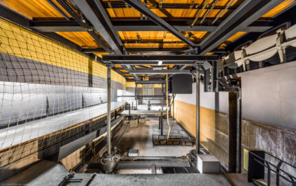 Schloss und Schlossgarten Schwetzingen - Garten - Schlosstheater - Von innen - Orchestergraben / Proszenium / Unterbühne - Von Süden - Blick von der südlichen Tür zum Orchestergraben nach Norden in den Raum unterhalb des Proszeniums; dieser Raum beherbergt eine Hebevorrichtung für das Anheben und Absenken des Bodens des Proszeniums; im Moment ist der Boden teilweise abgesenkt, daher sind die Türen im Moment nicht begehbar; oben im Bild die Unterseite des Bodens des Proszeniums, der auf einem Stahlgerüst mit Doppel-T Trägern liegt; unter dem Boden verlaufen Elektroleitungen zu Steckdosen für die Beleuchtung der Notenständer; links hinter dem weißen Netz der Raum unterhalb der Vorbühne, der zur Verlängerung des Orchestergrabens bei komplett abgesenktem Boden dient; rechts oben sind Stühle der Sitzreihen des Zuschauerraums zu sehen; links und rechts im Vordergrund und Hintergrund sind die vier Gewindespindeln mit den Antriebswellen und Getrieben zum Heben und Senken des Bodens des Proszeniums teilweise zu sehen (Gesamter Bau: Länge x Breite: ca. 62 x 35 Meter; Architekt: Nicolas de Pigage (1723-1796); Bauzeit: 1752-1753, Erweiterung 1762, Sanierung: 1937, Renovierung: 1952, Generalsanierung / Umbau 1971-1974, Restaurierung: 1999-2005) (aufgenommen im September 2025, am frühen Nachmittag) Schloss und Schlossgarten Schwetzingen - Garten - Schlosstheater - Von innen - Orchestergraben / Proszenium / Unterbühne - Von Süden - Blick von der südlichen Tür zum Orchestergraben nach Norden in den Raum unterhalb des Proszeniums; dieser Raum beherbergt eine Hebevorrichtung für das Anheben und Absenken des Bodens des Proszeniums; im Moment ist der Boden teilweise abgesenkt, daher sind die Türen im Moment nicht begehbar; oben im Bild die Unterseite des Bodens des Proszeniums, der auf einem Stahlgerüst mit Doppel-T Trägern liegt; unter dem Boden verlaufen Elektroleitungen zu Steckdosen für die Beleuchtung der Notenständer; links hinter dem weißen Netz der Raum unterhalb der Vorbühne, der zur Verlängerung des Orchestergrabens bei komplett abgesenktem Boden dient; rechts oben sind Stühle der Sitzreihen des Zuschauerraums zu sehen; links und rechts im Vordergrund und Hintergrund sind die vier Gewindespindeln mit den Antriebswellen und Getrieben zum Heben und Senken des Bodens des Proszeniums teilweise zu sehen (Gesamter Bau: Länge x Breite: ca. 62 x 35 Meter; Architekt: Nicolas de Pigage (1723-1796); Bauzeit: 1752-1753, Erweiterung 1762, Sanierung: 1937, Renovierung: 1952, Generalsanierung / Umbau 1971-1974, Restaurierung: 1999-2005) (aufgenommen im September 2025, am frühen Nachmittag)