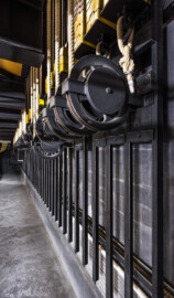 Schloss und Schlossgarten Schwetzingen - Garten - Schlosstheater - Von innen - Bühnenraum - Unterbühne - Von Nordwest - Blick von der Südwestecke der Unterbühne auf den unteren Teil der Südwand und auf die unteren Umlenkrollen der Handkonterzüge und elekrischen Züge; die dicken Hanfseile sind die Kommandoseile, mit denen die Zugstangen (beladen z.B. mit Bühnenprospekt oder Scheinwerfern) manuell in der Höhe verstellt werden können; je Zug sind vier Stahlseile mit Gegengewichtsschlitten verbunden, die über Umlenkrollen mit Zugstangen unterhalb des Rollenbodens verbunden sind; links oben sind Stahlträger und Platten des Bodens der Hauptbühne zu sehen (Gesamter Bau: Länge x Breite: ca. 62 x 35 Meter; Architekt: Nicolas de Pigage (1723-1796); Bauzeit: 1752-1753, Erweiterung 1762, Sanierung: 1937, Renovierung: 1952, Generalsanierung / Umbau 1971-1974, Restaurierung: 1999-2005) (aufgenommen im September 2025, am frühen Nachmittag) Schloss und Schlossgarten Schwetzingen - Garten - Schlosstheater - Von innen - Bühnenraum - Unterbühne - Von Nordwest - Blick von der Südwestecke der Unterbühne auf den unteren Teil der Südwand und auf die unteren Umlenkrollen der Handkonterzüge und elekrischen Züge; die dicken Hanfseile sind die Kommandoseile, mit denen die Zugstangen (beladen z.B. mit Bühnenprospekt oder Scheinwerfern) manuell in der Höhe verstellt werden können; je Zug sind vier Stahlseile mit Gegengewichtsschlitten verbunden, die über Umlenkrollen mit Zugstangen unterhalb des Rollenbodens verbunden sind; links oben sind Stahlträger und Platten des Bodens der Hauptbühne zu sehen (Gesamter Bau: Länge x Breite: ca. 62 x 35 Meter; Architekt: Nicolas de Pigage (1723-1796); Bauzeit: 1752-1753, Erweiterung 1762, Sanierung: 1937, Renovierung: 1952, Generalsanierung / Umbau 1971-1974, Restaurierung: 1999-2005) (aufgenommen im September 2025, am frühen Nachmittag)