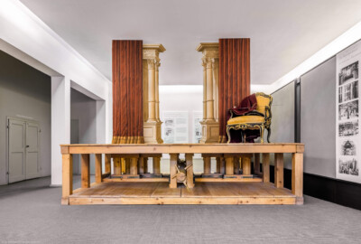 Schloss und Schlossgarten Schwetzingen - Garten - Schlosstheater - Von innen - 2. Obergeschoss (Rang), nördliches Foyer - Von Osten - Blick auf das Modell einer barocken Kulissenbühne aus Zuschauersicht; eine solche Kulissenbühne ist in Gassen eingeteilt, die von versetzt hintereinander angeordneten, meist bemalten Seitenkulissen gebildet werden; sie ermöglichen Auftritte von den Seiten und verbergen die Beleuchtung; die Seitenkulissen sind an vertikalen Hölzern befestigt, diese sind wiederum in der Unterbühne auf einem beweglichen horizontalen Schlitten, dem sog. Kulissenwagen, montiert; jeder Wagen ist mittels Schnurzügen mit einer zentralen Walze, dem sog. Wellenbaum, verbunden; dreht man den Wellenbaum, so können gleichzeitig mehrere Kulissenwagen verschoben werden; der Zuschauer sieht dadurch das gleichzeitige Erscheinen bzw. Verschwinden mehrerer Seitenkulissen (Gesamter Bau: Länge x Breite: ca. 62 x 35 Meter; Architekt: Nicolas de Pigage (1723-1796); Bauzeit: 1752-1753, Erweiterung 1762, Sanierung: 1937, Renovierung: 1952, Generalsanierung / Umbau 1971-1974, Restaurierung: 1999-2005) (aufgenommen im September 2025, am frühen Nachmittag)
