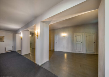 Schloss und Schlossgarten Schwetzingen - Garten - Schlosstheater - Von innen - Erdgeschoss, südliches Foyer - Von Osten - Blick von der Eingangstür zum südlichen Foyer nach Westen in das südliche Foyer; die Eingangstür ist geöffnet, sodass Sonnenlicht in das Foyer gelangen kann; urprünglich befand sich die südliche Mauer des Theaters an der Stelle, an der heute die zwei Pfeiler stehen; beim großen Umbau 1971-1974 wurde das Schlosstheater zum nördlichen Zirkelbau (nach Süden / links) hin erweitert; originaler Bau und Anbau erkennbar am Boden: Holzboden bedeutet originaler Bau, Teppichboden bedeutet Anbau des 20. Jahrhunderts; hinter den Pfeilern die Zugänge zu den Logen im Parterre (Gesamter Bau: Länge x Breite: ca. 62 x 35 Meter; Architekt: Nicolas de Pigage (1723-1796); Bauzeit: 1752-1753, Erweiterung 1762, Sanierung: 1937, Renovierung: 1952, Generalsanierung / Umbau 1971-1974, Restaurierung: 1999-2005) (aufgenommen im September 2025, am späten Vormittag)