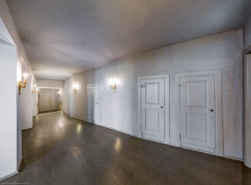 Schloss und Schlossgarten Schwetzingen - Garten - Schlosstheater - Von innen - Erdgeschoss, südliches Foyer - Von Osten - Blick von der Ostwand des Foyers nach Westen in das südliche Foyer; die Eingangstür zum südlichen Foyer (ganz links, nicht sichtbar) ist geöffnet, sodass Sonnenlicht in das Foyer gelangt; urprünglich befand sich die südliche Mauer des Theaters an der Stelle, an der heute die zwei Pfeiler stehen; beim großen Umbau 1971-1974 wurde das Schlosstheater zum nördlichen Zirkelbau (nach Süden / links) hin erweitert; originaler Bau und Anbau erkennbar am Boden: Holzboden bedeutet originaler Bau, Teppichboden bedeutet Anbau des 20. Jahrhunderts; rechts die Zugänge zu den Logen im Parterre; die Tür links im Hintergrund führt zur Bühne, dem Orchestergraben und der Unterbühne; die (nicht sichtbare) Tür rechts davon führt in das Parterre des Zuschauerraums; der Durchgang ganz rechts (beim Türrahmen) führt in das östliche Foyer (Gesamter Bau: Länge x Breite: ca. 62 x 35 Meter; Architekt: Nicolas de Pigage (1723-1796); Bauzeit: 1752-1753, Erweiterung 1762, Sanierung: 1937, Renovierung: 1952, Generalsanierung / Umbau 1971-1974, Restaurierung: 1999-2005) (aufgenommen im September 2025, um die Mittagszeit)