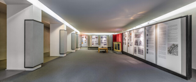 Schloss und Schlossgarten Schwetzingen - Garten - Schlosstheater - Von innen - Erdgeschoss, nördliches Foyer - Von Osten - Blick nach Westen in das nördliche Foyer im Erdgeschoss;  urprünglich befand sich die nördliche Mauer des Theaters an der Stelle, an der heute die drei Pfeiler stehen; in den 1950/1960er Jahren und vor allem beim großen Umbau 1971-1974 wurde das Schlosstheater nach Norden hin erweitert; originaler Bau und Anbau erkennbar am Boden: Holzboden bedeutet originaler Bau, Teppichboden bedeutet Anbau des 20. Jahrhunderts; hinten und rechts Tafeln einer Ausstellung zur Geschichte des Theaters; rechts unterhalb der Bildmitte eine Vitrine mit Kulturmüllfunden, geborgen aus z.B. Hohlräumen oder Dachfußbereichen während der sanierungsbegleitenden bauhistorischen Untersuchung des Schwetzinger Hoftheaters durch Robert Erb und Peter Knoch, 1999-2005; rechts der Vitrine ein Nachbau eines Teils der historischen Brüstung zwischen dem Parterre und dem Orchesterraum (Gesamter Bau: Länge x Breite: ca. 62 x 35 Meter; Architekt: Nicolas de Pigage (1723-1796); Bauzeit: 1752-1753, Erweiterung 1762, Sanierung: 1937, Renovierung: 1952, Generalsanierung / Umbau 1971-1974, Restaurierung: 1999-2005) (aufgenommen im September 2025, am späten Vormittag)