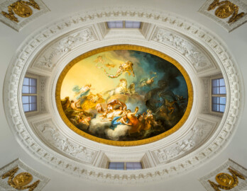 Schloss und Schlossgarten Schwetzingen - Garten - Badhaus - Von innen - Ovalsaal - Deckengemälde - Von unten - Blick nach oben in den Tambour und auf das Deckengemälde "Aurora vertreibt die Nacht"; links Aurora (Göttin der Morgenröte, mit blauem Mantel, auf goldenem Wagen, von zwei weißen Sonnenpferden gezogen), darüber schwebende Putti mit Rosenkränzen, links unten Uranus (Gott des Himmels und Urahn der Götter, versucht Aurora zurück zu halten), links ganz unten zwei spielende Putti mit Jagdhorn, unterhalb Bildmitte Tauschwestern (auf Wolkenbank, aus goldenen Gefäßen den Tau spendend), rechts etwas oberhalb der Bildmitte der Morgenstern (mit Fackel, Aurora den Weg zeigend), ganz rechts eine Eule (als Nachtvogel, den Weg zeigend) und Fledermäuse, links davon Nox (die mohnbekrönte Göttin der Nacht) (Maler: Nicolas Guibal (1725-1784); Datierung: 1772; Signaturen: links unten "N. Guibal Lotharingus inv et fecit 1772" und rechts unten "N. Guibal Lotharingus invenit et pinxit 1772"; Öl auf Leinwand, mit vergoldeten Eichenlaubkranz eingefasst; Restaurierungen: 1890, 1949, 2006) (Gesamter Bau: Länge x Breite ca. 18 x 14 Meter, Höhe ca. 10 Meter; Architekt: Nicolas de Pigage (1723-1796); Bauzeit: 1768-1772; Gesamtinstandsetzung innen und außen: 1999-2006) (aufgenommen im Oktober 2025, am frühen Nachmittag) Schloss und Schlossgarten Schwetzingen - Garten - Badhaus - Von innen - Ovalsaal - Deckengemälde - Von unten - Blick nach oben in den Tambour und auf das Deckengemälde "Aurora vertreibt die Nacht"; links Aurora (Göttin der Morgenröte, mit blauem Mantel, auf goldenem Wagen, von zwei weißen Sonnenpferden gezogen), darüber schwebende Putti mit Rosenkränzen, links unten Uranus (Gott des Himmels und Urahn der Götter, versucht Aurora zurück zu halten), links ganz unten zwei spielende Putti mit Jagdhorn, unterhalb Bildmitte Tauschwestern (auf Wolkenbank, aus goldenen Gefäßen den Tau spendend), rechts etwas oberhalb der Bildmitte der Morgenstern (mit Fackel, Aurora den Weg zeigend), ganz rechts eine Eule (als Nachtvogel, den Weg zeigend) und Fledermäuse, links davon Nox (die mohnbekrönte Göttin der Nacht) (Maler: Nicolas Guibal (1725-1784); Datierung: 1772; Signaturen: links unten "N. Guibal Lotharingus inv et fecit 1772" und rechts unten "N. Guibal Lotharingus invenit et pinxit 1772"; Öl auf Leinwand, mit vergoldeten Eichenlaubkranz eingefasst; Restaurierungen: 1890, 1949, 2006) (Gesamter Bau: Länge x Breite ca. 18 x 14 Meter, Höhe ca. 10 Meter; Architekt: Nicolas de Pigage (1723-1796); Bauzeit: 1768-1772; Gesamtinstandsetzung innen und außen: 1999-2006) (aufgenommen im Oktober 2025, am frühen Nachmittag)