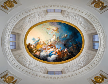 Schloss und Schlossgarten Schwetzingen - Garten - Badhaus - Von innen - Ovalsaal - Deckengemälde - Von unten - Blick nach oben in den Tambour und auf das Deckengemälde "Aurora vertreibt die Nacht"; links Aurora (Göttin der Morgenröte, mit blauem Mantel, auf goldenem Wagen, von zwei weißen Sonnenpferden gezogen), darüber schwebende Putti mit Rosenkränzen, links unten Uranus (Gott des Himmels und Urahn der Götter, versucht Aurora zurück zu halten), links ganz unten zwei spielende Putti mit Jagdhorn, unterhalb Bildmitte Tauschwestern (auf Wolkenbank, aus goldenen Gefäßen den Tau spendend), rechts etwas oberhalb der Bildmitte der Morgenstern (mit Fackel, Aurora den Weg zeigend), ganz rechts eine Eule (als Nachtvogel, den Weg zeigend) (Maler: Nicolas Guibal (1725-1784); Datierung: 1772; Signaturen: links unten "N. Guibal Lotharingus inv et fecit 1772" und rechts unten "N. Guibal Lotharingus invenit et pinxit 1772"; Öl auf Leinwand, mit vergoldeten Eichenlaubkranz eingefasst; Restaurierungen: 1890, 1949, 2006) (Gesamter Bau: Länge x Breite ca. 18 x 15 Meter, Höhe ca. 11 Meter; Architekt: Nicolas de Pigage (1723-1796); Bauzeit: 1768-1772; Gesamtinstandsetzung innen und außen: 1999-2006) (aufgenommen im Oktober 2025, am frühen Nachmittag) Schloss und Schlossgarten Schwetzingen - Garten - Badhaus - Von innen - Ovalsaal - Deckengemälde - Von unten - Blick nach oben in den Tambour und auf das Deckengemälde "Aurora vertreibt die Nacht"; links Aurora (Göttin der Morgenröte, mit blauem Mantel, auf goldenem Wagen, von zwei weißen Sonnenpferden gezogen), darüber schwebende Putti mit Rosenkränzen, links unten Uranus (Gott des Himmels und Urahn der Götter, versucht Aurora zurück zu halten), links ganz unten zwei spielende Putti mit Jagdhorn, unterhalb Bildmitte Tauschwestern (auf Wolkenbank, aus goldenen Gefäßen den Tau spendend), rechts etwas oberhalb der Bildmitte der Morgenstern (mit Fackel, Aurora den Weg zeigend), ganz rechts eine Eule (als Nachtvogel, den Weg zeigend) (Maler: Nicolas Guibal (1725-1784); Datierung: 1772; Signaturen: links unten "N. Guibal Lotharingus inv et fecit 1772" und rechts unten "N. Guibal Lotharingus invenit et pinxit 1772"; Öl auf Leinwand, mit vergoldeten Eichenlaubkranz eingefasst; Restaurierungen: 1890, 1949, 2006) (Gesamter Bau: Länge x Breite ca. 18 x 15 Meter, Höhe ca. 11 Meter; Architekt: Nicolas de Pigage (1723-1796); Bauzeit: 1768-1772; Gesamtinstandsetzung innen und außen: 1999-2006) (aufgenommen im Oktober 2025, am frühen Nachmittag)