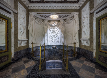 Schloss und Schlossgarten Schwetzingen - Garten - Badhaus - Von innen - Baderaum - Von Westen - Blick nach Osten in das Badezimmer, als Grotte gestaltet; unten in der Mitte sieben Stufen, das ovale Wasserbecken, eingefasst von einem Metallgitter, darüber Wasserspender (zentrale Messingurne und metallene Schlangen), darüber ein großer Stuckvorhang, darüber eine runde Öffnung zur Beleuchtung und Belüftung, ganz links eine Tür nach außen, ganz rechts eine Tür zum westlichen Vorzimmer, links und rechts zwei (von sechs) Wandfelder mit Reliefdarstellungen von Najaden (Nymphen der griechischen Mythologie) (Stuckateur: Giuseppe Antonio Pozzi (1732–1811)) (Gesamter Bau: Länge x Breite ca. 18 x 14 Meter, Höhe ca. 10 Meter; Architekt: Nicolas de Pigage (1723-1796); Bauzeit: 1768-1772; Gesamtinstandsetzung innen und außen: 1999-2006) (aufgenommen im Oktober 2025, am späten Nachmittag) Schloss und Schlossgarten Schwetzingen - Garten - Badhaus - Von innen - Baderaum - Von Westen - Blick nach Osten in das Badezimmer, als Grotte gestaltet; unten in der Mitte sieben Stufen, das ovale Wasserbecken, eingefasst von einem Metallgitter, darüber Wasserspender (zentrale Messingurne und metallene Schlangen), darüber ein großer Stuckvorhang, darüber eine runde Öffnung zur Beleuchtung und Belüftung, ganz links eine Tür nach außen, ganz rechts eine Tür zum westlichen Vorzimmer, links und rechts zwei (von sechs) Wandfelder mit Reliefdarstellungen von Najaden (Nymphen der griechischen Mythologie) (Stuckateur: Giuseppe Antonio Pozzi (1732–1811)) (Gesamter Bau: Länge x Breite ca. 18 x 14 Meter, Höhe ca. 10 Meter; Architekt: Nicolas de Pigage (1723-1796); Bauzeit: 1768-1772; Gesamtinstandsetzung innen und außen: 1999-2006) (aufgenommen im Oktober 2025, am späten Nachmittag)