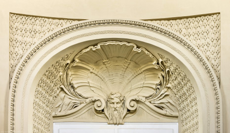 Schloss und Schlossgarten Schwetzingen - Garten - Badhaus - Von innen - Ovalsaal - Südliche Eingangstür - Von Norden - Blick von der Mitte des Ovalsaals nach Süden auf die Rundbogennische der südlichen Tür; Tropfenstuck im Feld über dem Rundbogen, ebenso im Gewände der Tür; auf dem Rundbogen ein Blattstab, Perl- und Eierstab; im Bogenfeld ein bärtiger Neptunskopf mit Schilf im Haar, hinterfangen von einer großen, stuckierten Muschel, flankiert von Schilfrohrblättern; beiderseits des Kopfes Voluten (Stuckateur: Giuseppe Antonio Pozzi (1732–1811) zugeschrieben) (Gesamter Bau: Länge x Breite ca. 18 x 14 Meter, Höhe ca. 10 Meter; Architekt: Nicolas de Pigage (1723-1796); Bauzeit: 1768-1772; Gesamtinstandsetzung innen und außen: 1999-2006) (aufgenommen im Oktober 2025, am frühen Nachmittag) Schloss und Schlossgarten Schwetzingen - Garten - Badhaus - Von innen - Ovalsaal - Südliche Eingangstür - Von Norden - Blick von der Mitte des Ovalsaals nach Süden auf die Rundbogennische der südlichen Tür; Tropfenstuck im Feld über dem Rundbogen, ebenso im Gewände der Tür; auf dem Rundbogen ein Blattstab, Perl- und Eierstab; im Bogenfeld ein bärtiger Neptunskopf mit Schilf im Haar, hinterfangen von einer großen, stuckierten Muschel, flankiert von Schilfrohrblättern; beiderseits des Kopfes Voluten (Stuckateur: Giuseppe Antonio Pozzi (1732–1811) zugeschrieben) (Gesamter Bau: Länge x Breite ca. 18 x 14 Meter, Höhe ca. 10 Meter; Architekt: Nicolas de Pigage (1723-1796); Bauzeit: 1768-1772; Gesamtinstandsetzung innen und außen: 1999-2006) (aufgenommen im Oktober 2025, am frühen Nachmittag)