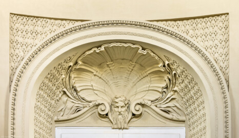 Schloss und Schlossgarten Schwetzingen - Garten - Badhaus - Von innen - Ovalsaal - Südliche Eingangstür - Von Norden - Blick von der Mitte des Ovalsaals nach Süden auf die Rundbogennische der südlichen Tür; Tropfenstuck im Feld über dem Rundbogen, ebenso im Gewände der Tür; auf dem Rundbogen ein Blattstab, Perl- und Eierstab; im Bogenfeld ein bärtiger Neptunskopf mit Schilf im Haar, hinterfangen von einer großen, stuckierten Muschel, flankiert von Schilfrohrblättern; beiderseits des Kopfes Voluten (Stuckateur: Giuseppe Antonio Pozzi (1732–1811) zugeschrieben) (Gesamter Bau: Länge x Breite ca. 18 x 14 Meter, Höhe ca. 10 Meter; Architekt: Nicolas de Pigage (1723-1796); Bauzeit: 1768-1772; Gesamtinstandsetzung innen und außen: 1999-2006) (aufgenommen im Oktober 2025, am frühen Nachmittag) Schloss und Schlossgarten Schwetzingen - Garten - Badhaus - Von innen - Ovalsaal - Südliche Eingangstür - Von Norden - Blick von der Mitte des Ovalsaals nach Süden auf die Rundbogennische der südlichen Tür; Tropfenstuck im Feld über dem Rundbogen, ebenso im Gewände der Tür; auf dem Rundbogen ein Blattstab, Perl- und Eierstab; im Bogenfeld ein bärtiger Neptunskopf mit Schilf im Haar, hinterfangen von einer großen, stuckierten Muschel, flankiert von Schilfrohrblättern; beiderseits des Kopfes Voluten (Stuckateur: Giuseppe Antonio Pozzi (1732–1811) zugeschrieben) (Gesamter Bau: Länge x Breite ca. 18 x 14 Meter, Höhe ca. 10 Meter; Architekt: Nicolas de Pigage (1723-1796); Bauzeit: 1768-1772; Gesamtinstandsetzung innen und außen: 1999-2006) (aufgenommen im Oktober 2025, am frühen Nachmittag)